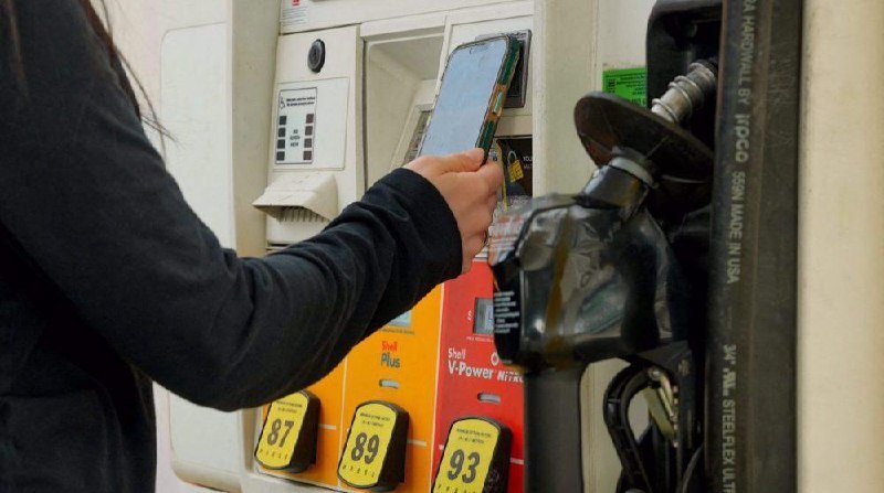 A person in a black long-sleeve shirt holds a smartphone near a Shell gas pump displaying fuel grade buttons labeled 87, 89, and 93.