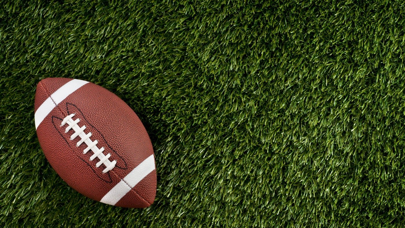 A brown American football with white laces rests on green artificial turf.