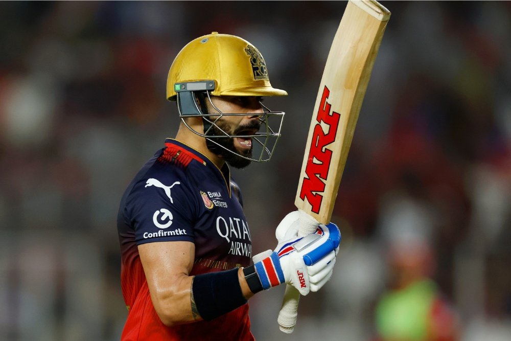 A bearded cricket batsman wearing a yellow helmet and dark blue jersey with red trim holds an MRF-branded bat upward against a blurred stadium crowd.