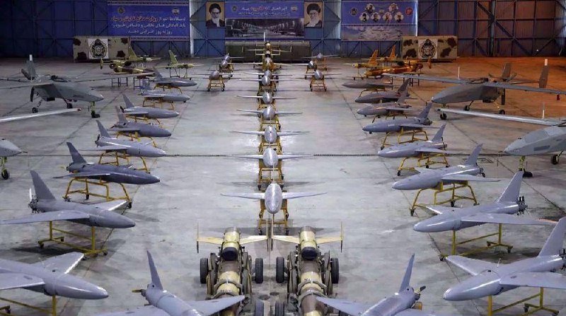 Rows of unmanned military aircraft and missiles are displayed on stands inside a hangar, with banners bearing portraits and text mounted on the back wall.