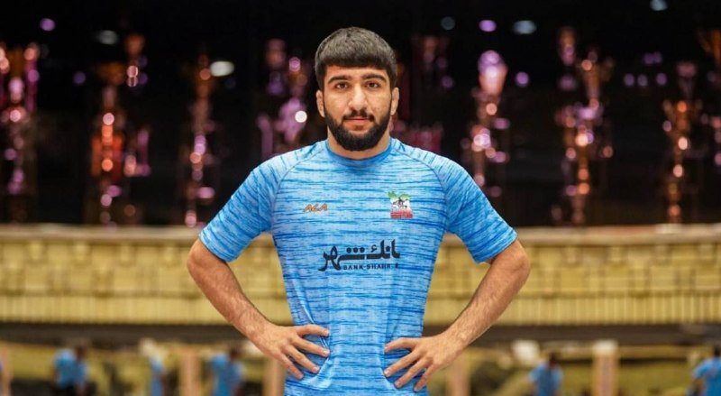 A bearded male athlete in a blue jersey stands with hands on hips in front of a row of trophies.