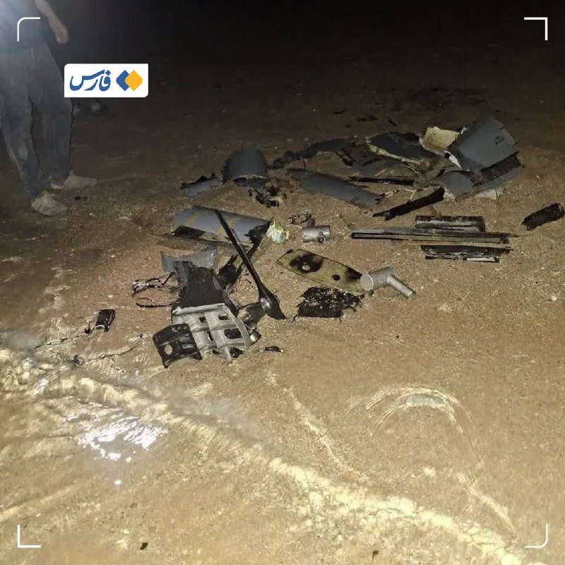 Scattered debris from a destroyed electronic device lies on a sandy surface, with a person's legs visible in the background.