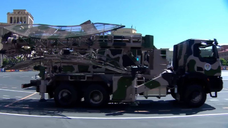 A camouflaged military truck carrying a large radar or satellite dish system is parked on a paved road with buildings in the background.