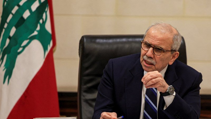 A bespectacled man with a mustache, wearing a dark suit and striped tie, speaks while seated at a desk beside a Lebanese flag.