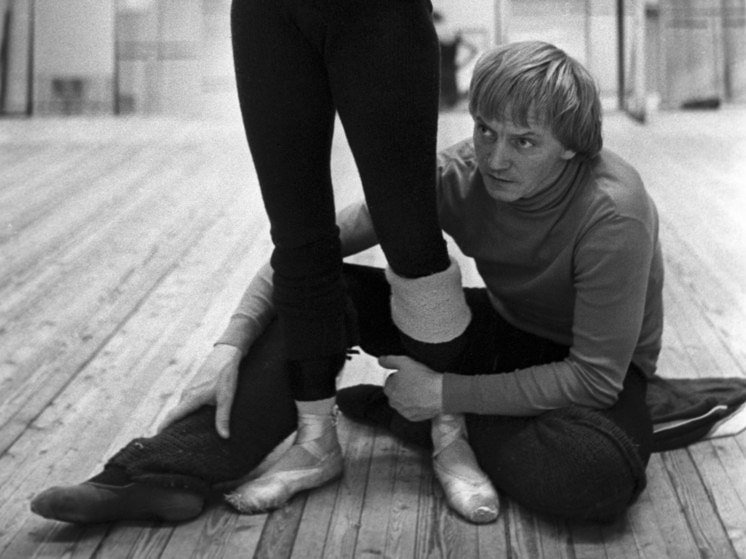 A man kneels on a wooden studio floor, adjusting another dancer's pointe shoe while a third dancer stands nearby.