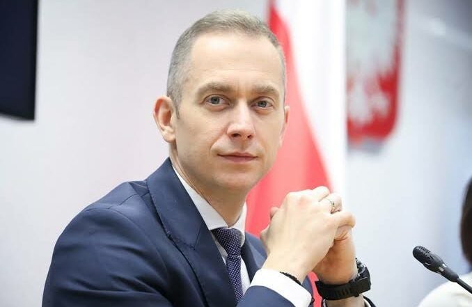 A man in a dark suit and tie sits at a table with his hands clasped, a microphone visible nearby and a red-and-white flag in the background.