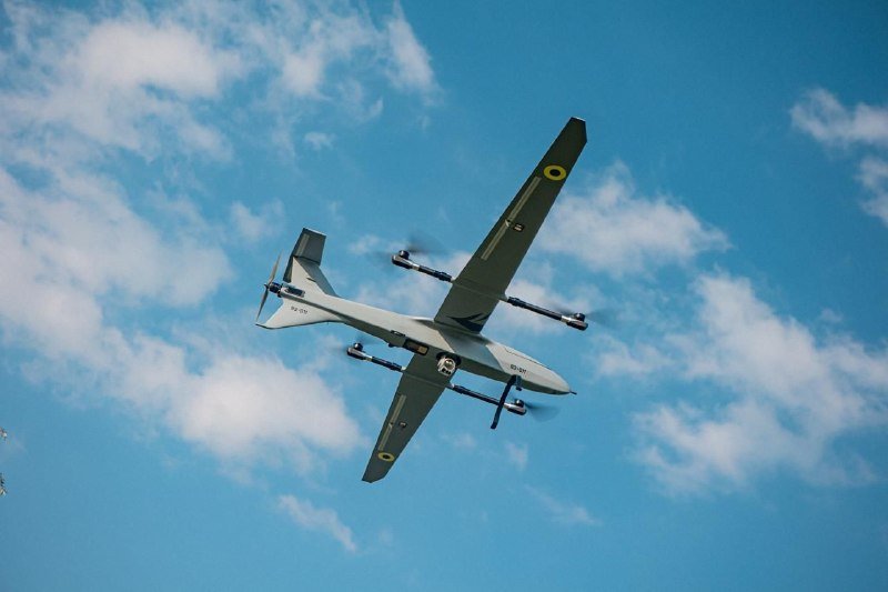 A grey military drone with four propellers flies through a partly cloudy blue sky, viewed from below.