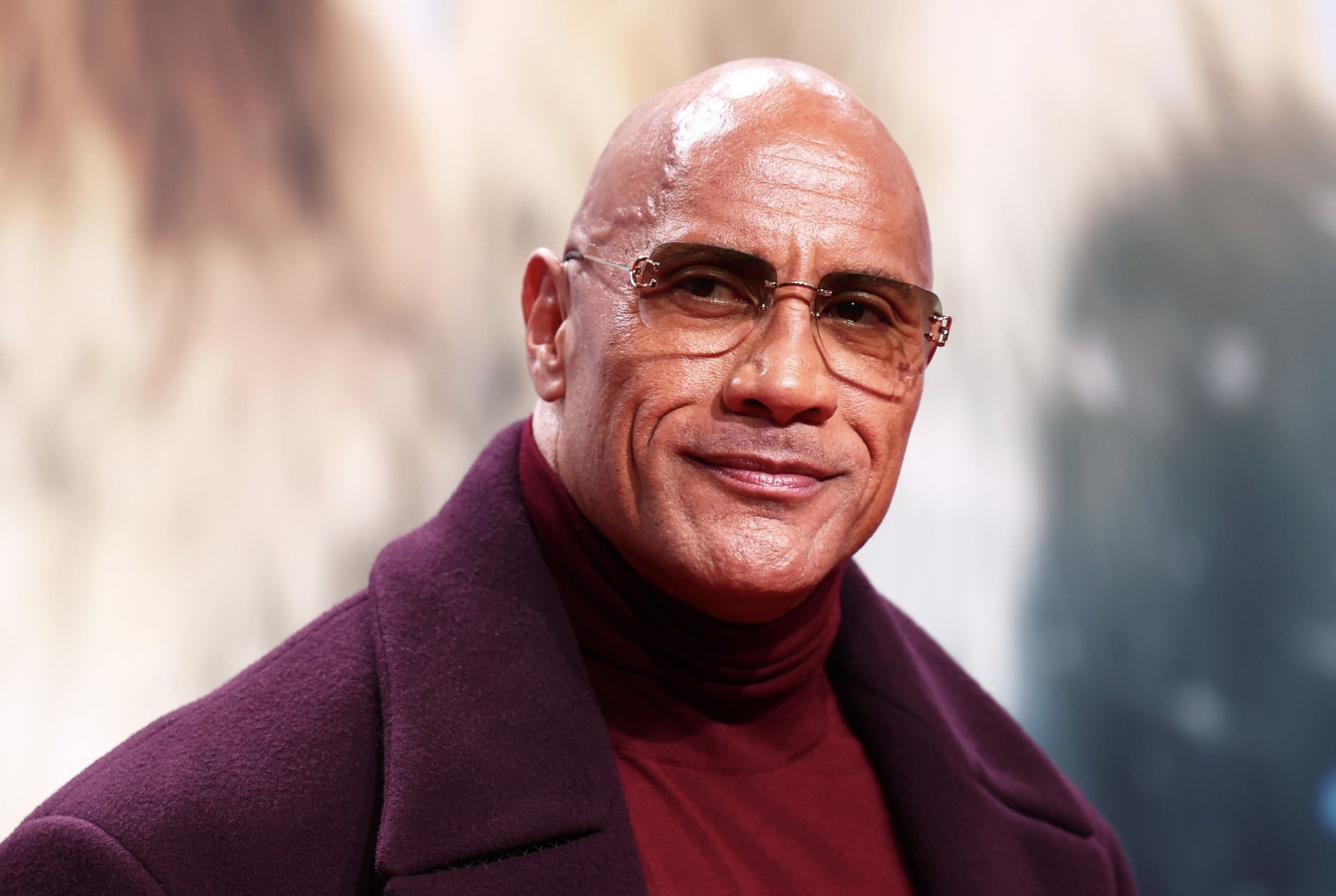 A bald man wearing rimless glasses, a burgundy turtleneck, and a purple coat smiles toward the camera.