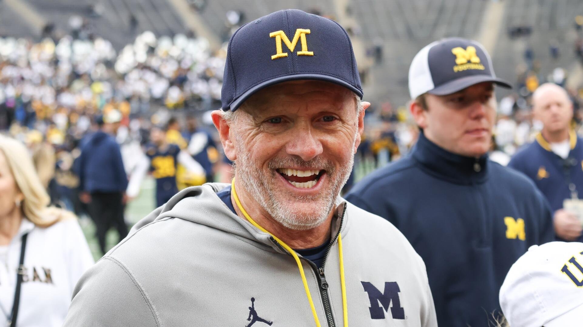 A smiling gray-bearded man wearing a navy "M" cap and gray hoodie stands on a football field, with a marching band and stadium crowd visible in the background.