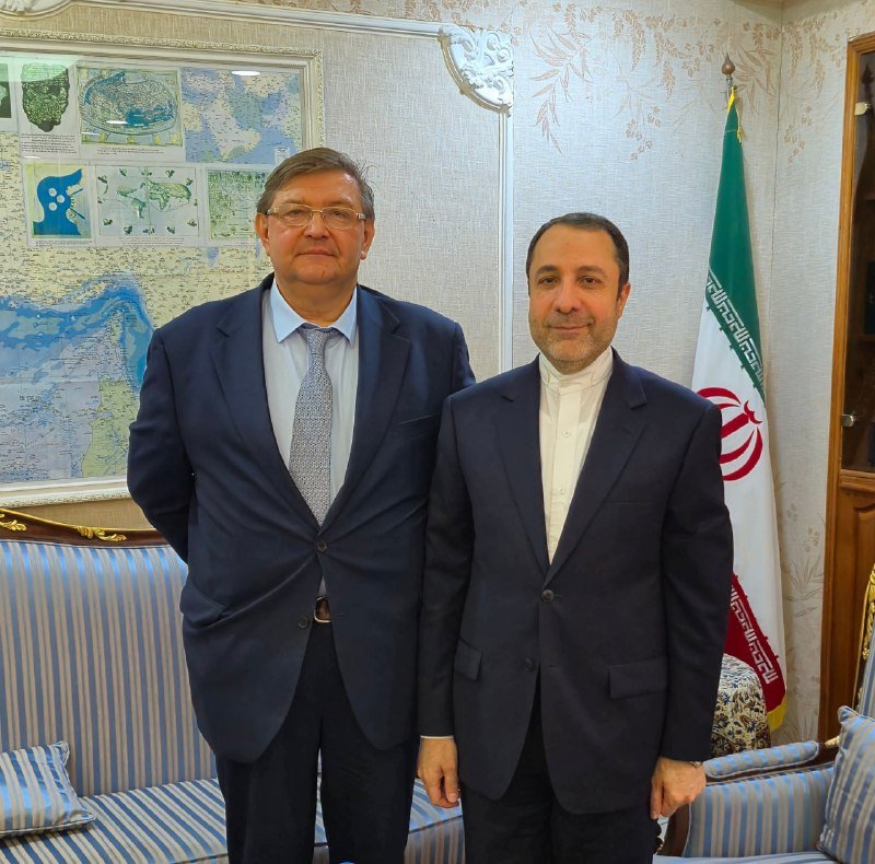 Two men in suits pose together indoors beside an Iranian flag and a framed historical map.