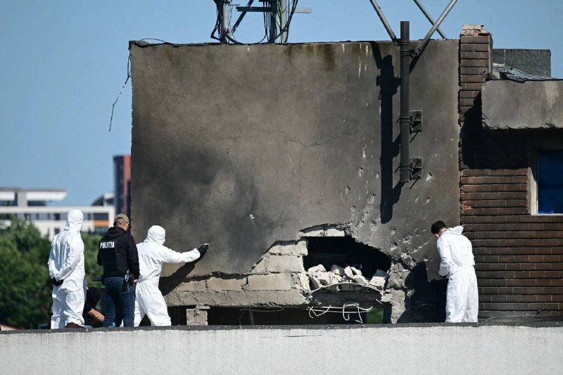 Investigators in white protective suits and one marked "POLITII" examine a damaged concrete wall with a large broken section on a rooftop.