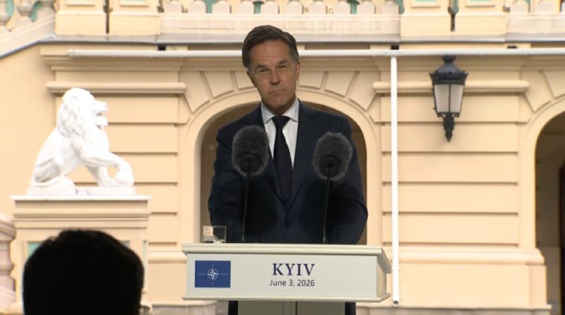 A man in a dark suit and tie stands at a podium labeled "KYIV June 3, 2026" with a NATO emblem, speaking behind two microphones outside a building with a stone lion statue.