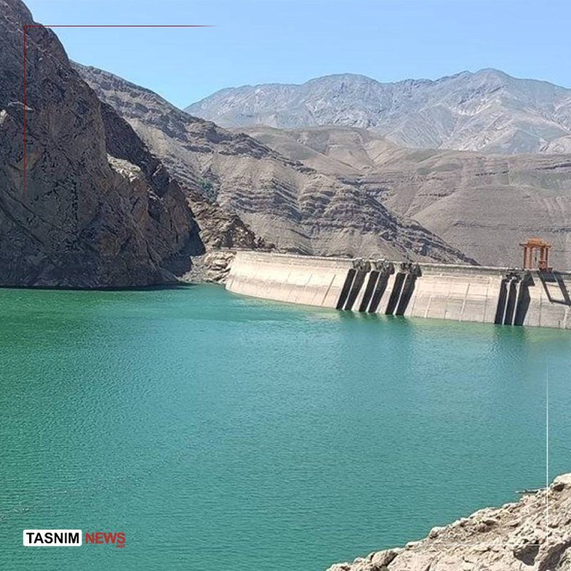 A concrete dam holds back turquoise reservoir water, set against barren, rugged mountains under a clear sky, with a "Tasnim News" logo in the corner.