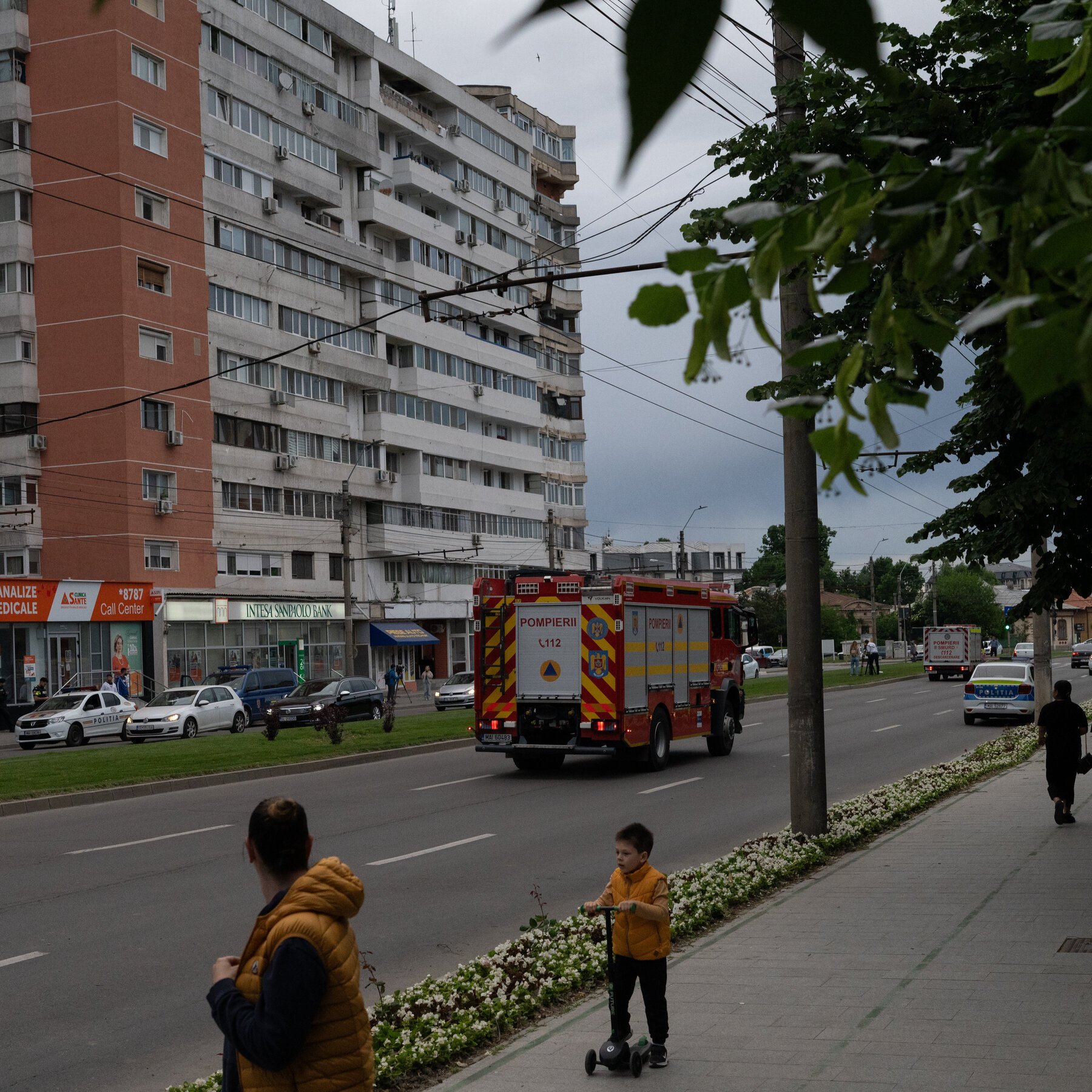 A fire truck and emergency vehicles travel down a city street lined with apartment buildings, while a child on a scooter watches from the sidewalk.
