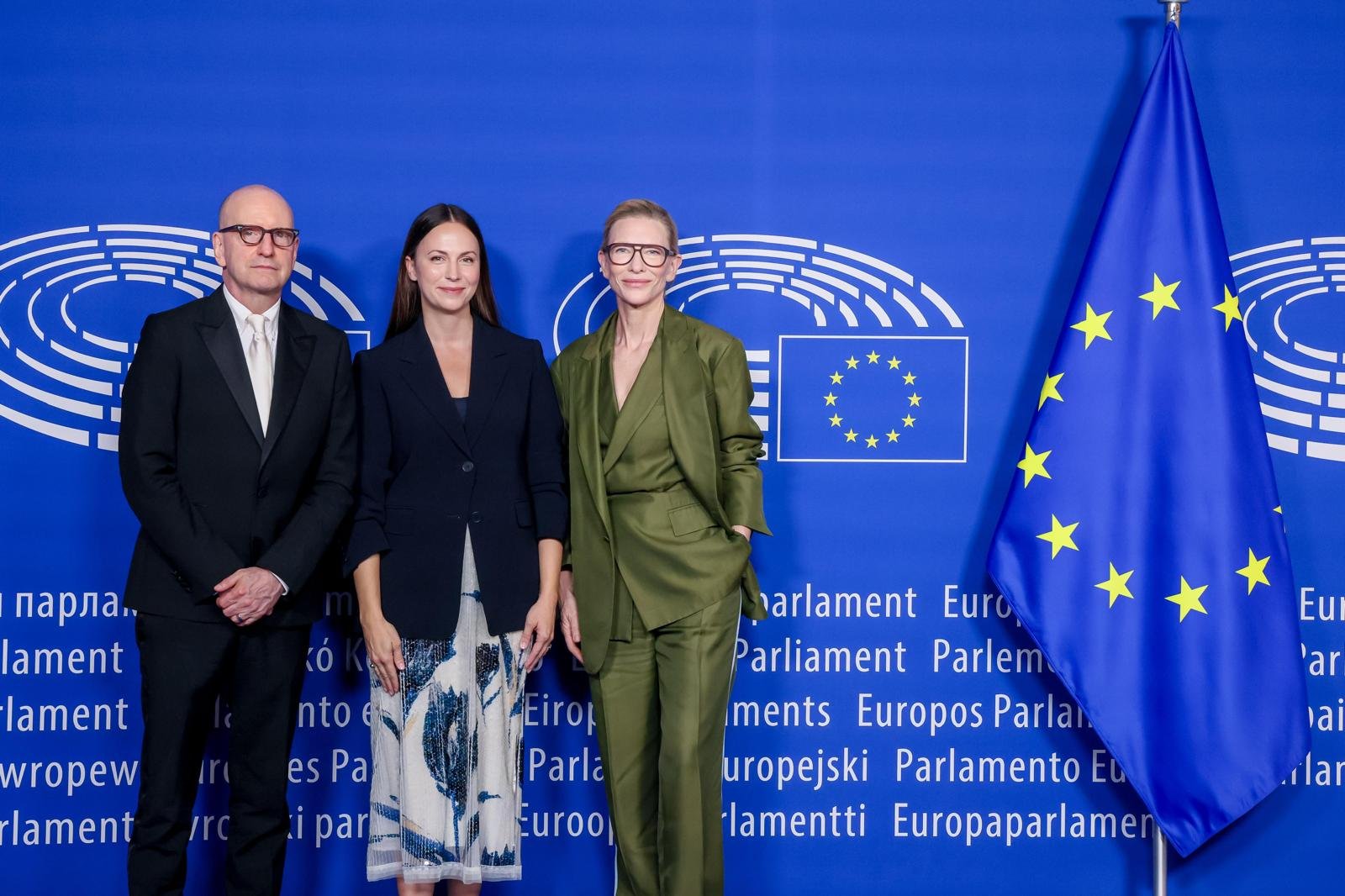 Three people pose in front of a blue backdrop displaying European Parliament logos in multiple languages, with an EU flag to the right.