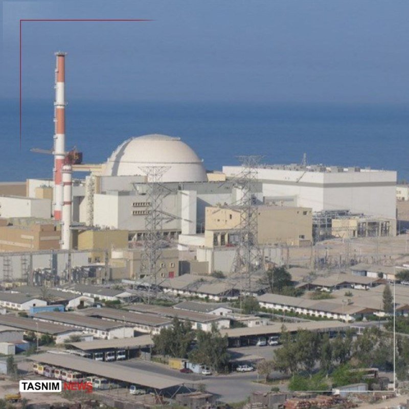 A coastal industrial facility with a domed containment building and a tall red-and-white striped smokestack sits beside the sea, with power transmission towers and low warehouse structures in the foreground.