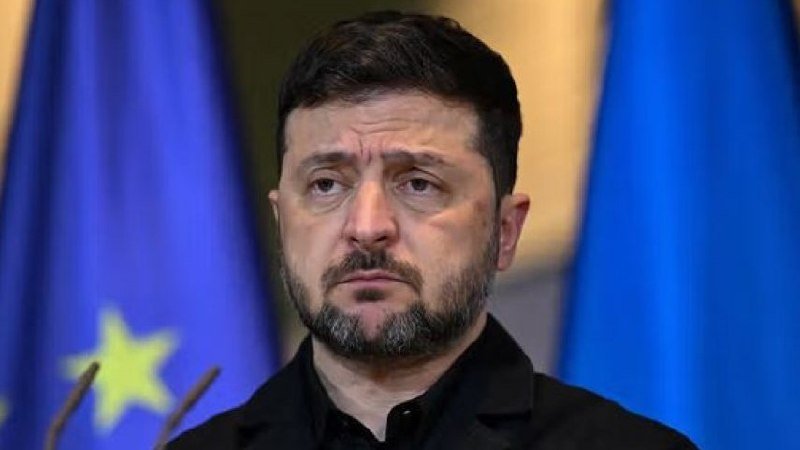 A bearded man in a black shirt stands before blue flags with yellow stars, looking somber near two microphones.
