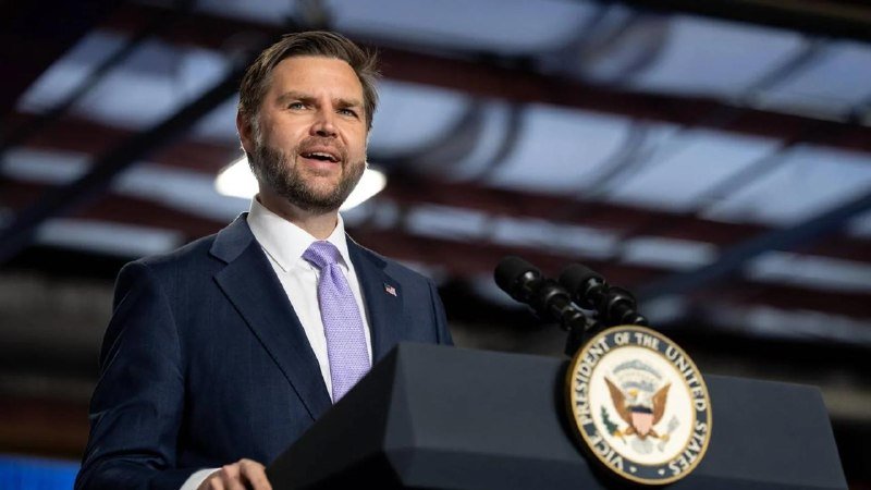 A bearded man in a dark blue suit and purple tie speaks at a podium bearing the Vice President of the United States seal.