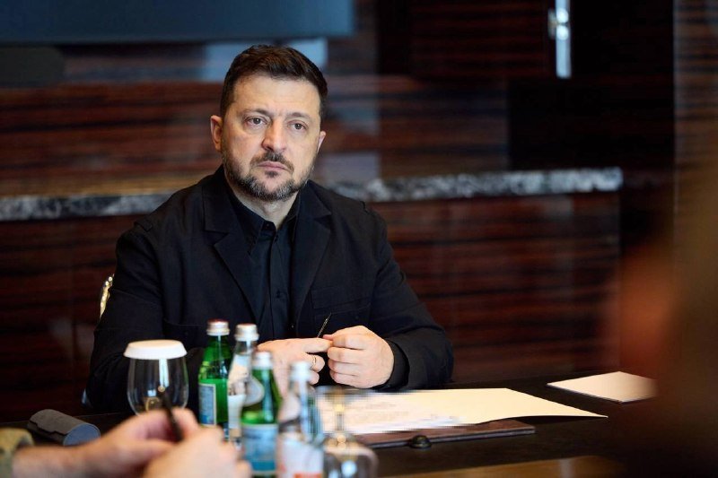 A bearded man in a black shirt and jacket sits at a table with water bottles and papers, holding a pen in a dimly lit room with wooden paneling.