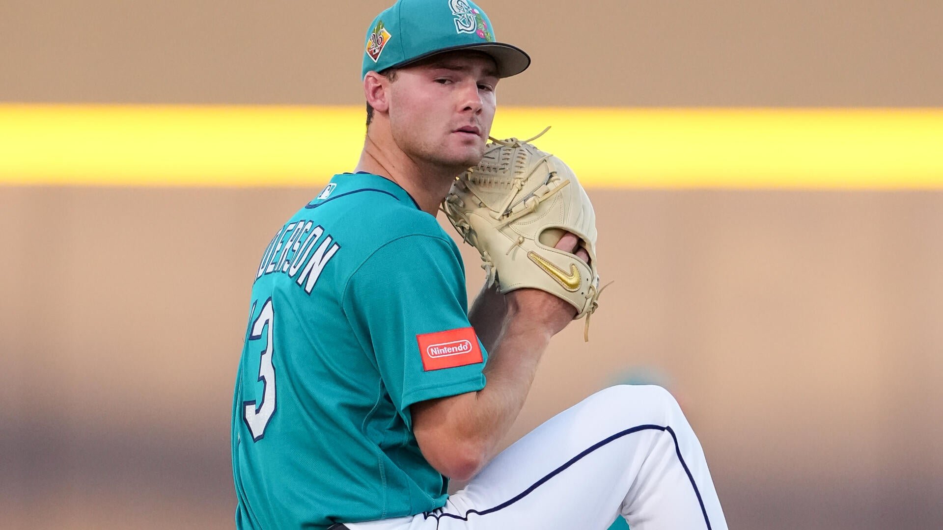 A baseball pitcher in a teal jersey winds up with a tan glove raised, mid-pitch on the mound.