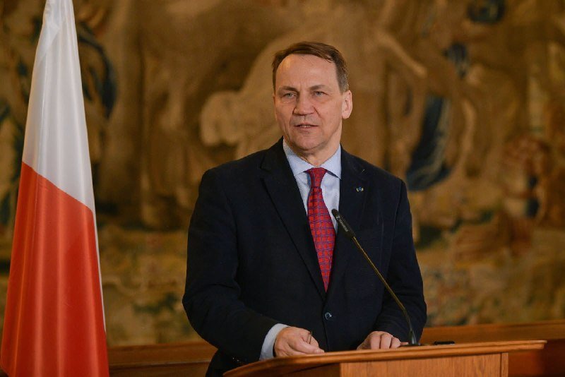 A man in a dark suit and red tie stands at a wooden podium beside a red-and-white flag, speaking into a microphone against a tapestry backdrop.