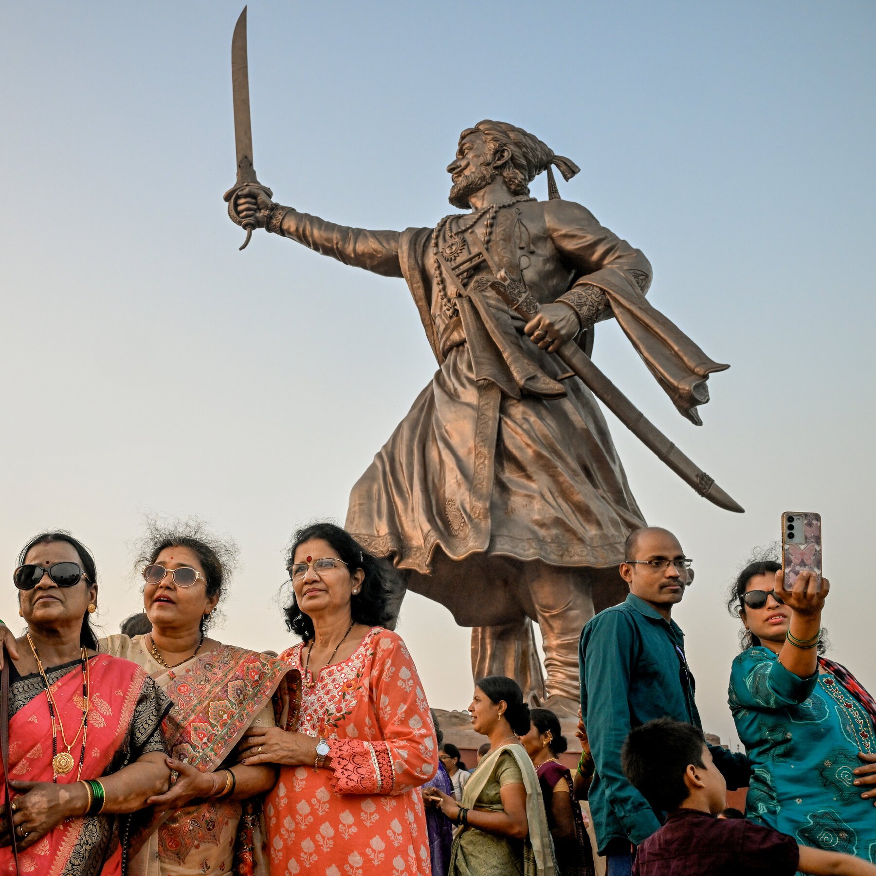 People in traditional Indian attire pose for a smartphone selfie in front of a large bronze statue of a sword-wielding warrior.