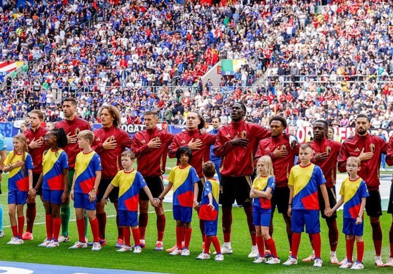 Soccer players in red jerseys stand with hands over their hearts alongside children in yellow, blue, and red uniforms on a field before a packed stadium crowd.