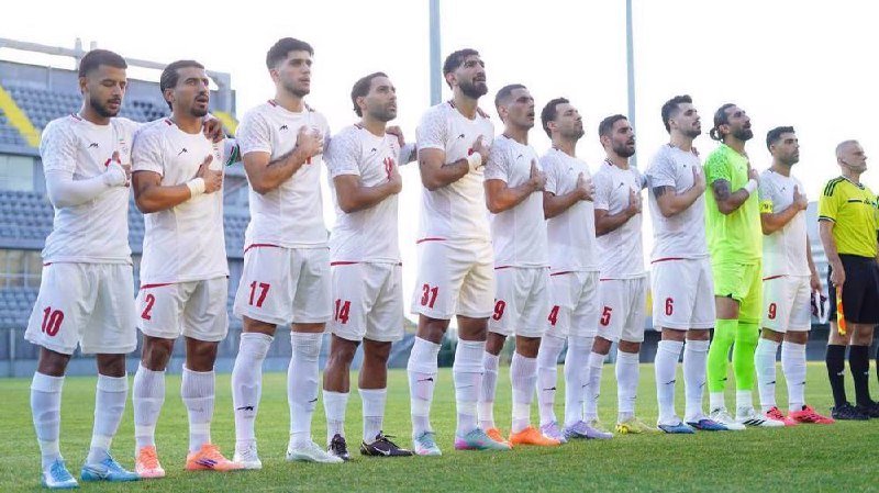 A soccer team in white and red uniforms stands in a line on a green field, hands over their hearts, alongside a goalkeeper in neon green and a referee.