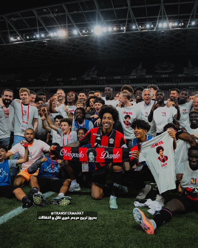 A large group of soccer players and staff celebrate on a stadium pitch at night, holding a "Obrigado Dante" banner and a printed t-shirt.