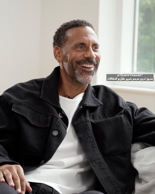 A smiling man with a graying beard, wearing a black denim jacket over a white shirt, sits indoors near a bright window during what appears to be an interview.