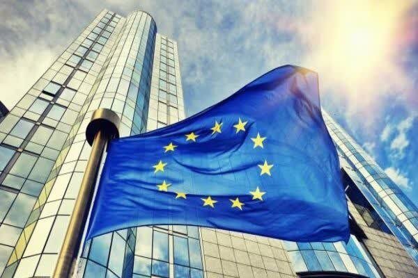 The European Union flag waves in the foreground against a backdrop of a tall, modern glass skyscraper under a partly cloudy sky.