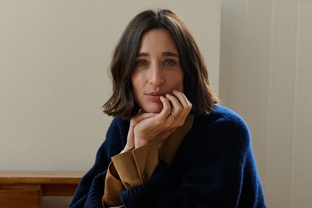 A woman with shoulder-length dark hair sits indoors against a plain beige wall, resting her chin on her hand while wearing a navy cardigan over a tan blouse.