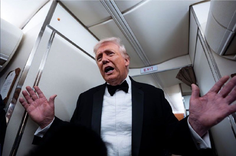 An older man with white hair wearing a black tuxedo and bow tie gestures with both hands raised while speaking inside what appears to be an aircraft cabin.