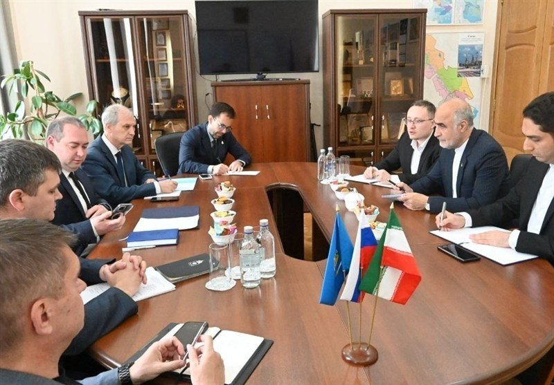 Men in suits sit around a wooden conference table with notepads, water bottles, and small flags, in an office featuring display cabinets, a wall-mounted screen, and a map.