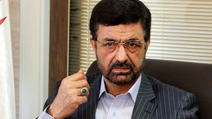 A bearded man in a pinstripe suit and glasses sits indoors with his fist raised, wearing a ring with a green stone.