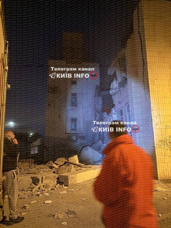 A nighttime scene shows a damaged building with collapsed walls and rubble scattered on the ground, as two people stand nearby observing the destruction.