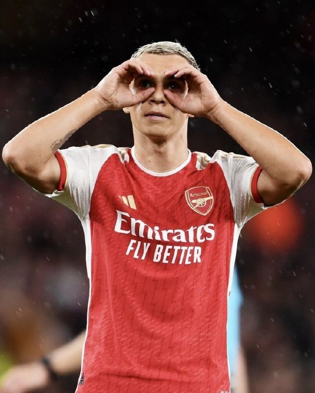 A soccer player in a red Arsenal jersey with "Emirates FLY BETTER" text makes binoculars with his hands near his eyes during a match.