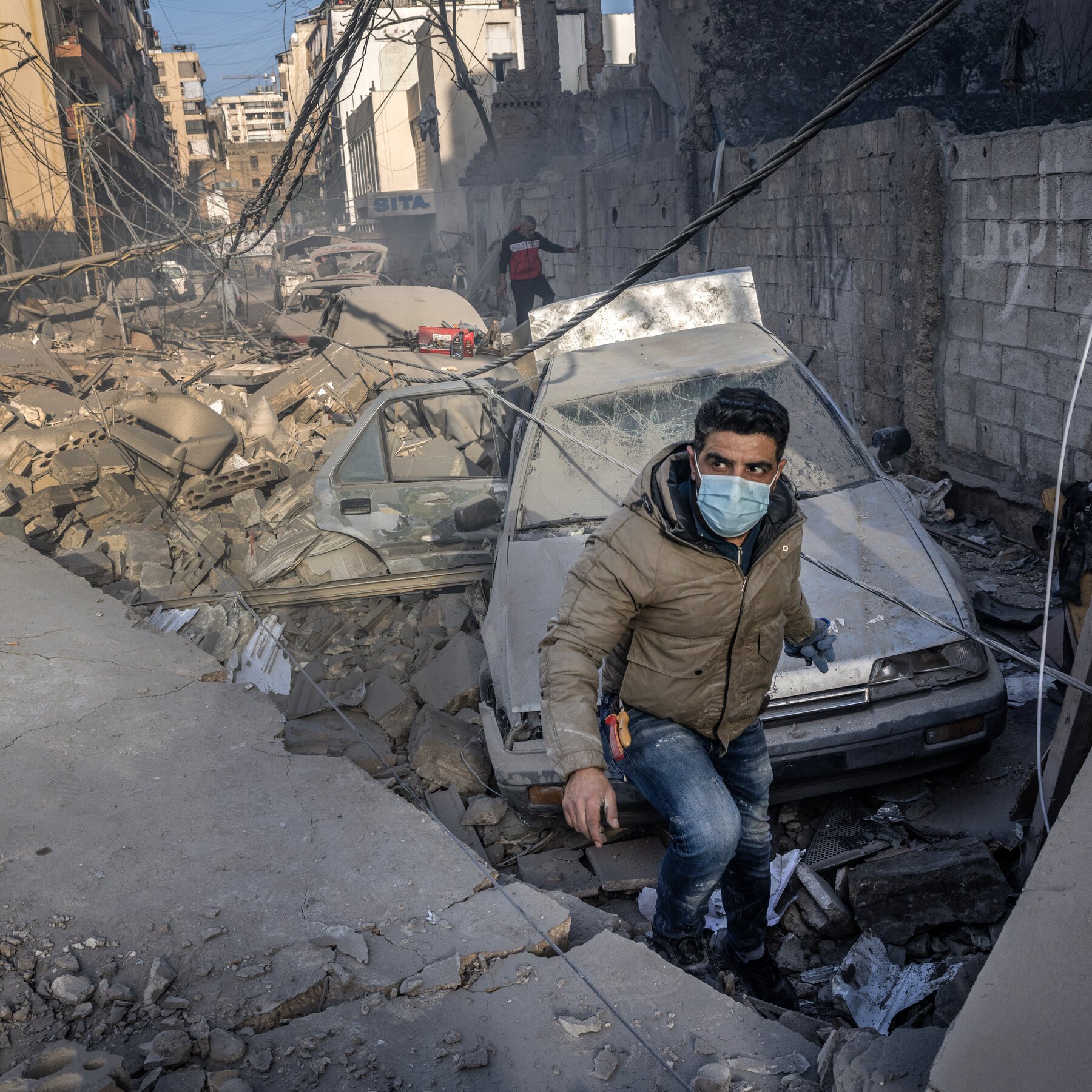 A masked man in a brown jacket steps through rubble and a crushed car on a debris-strewn street, with damaged buildings and tangled wires in the background.