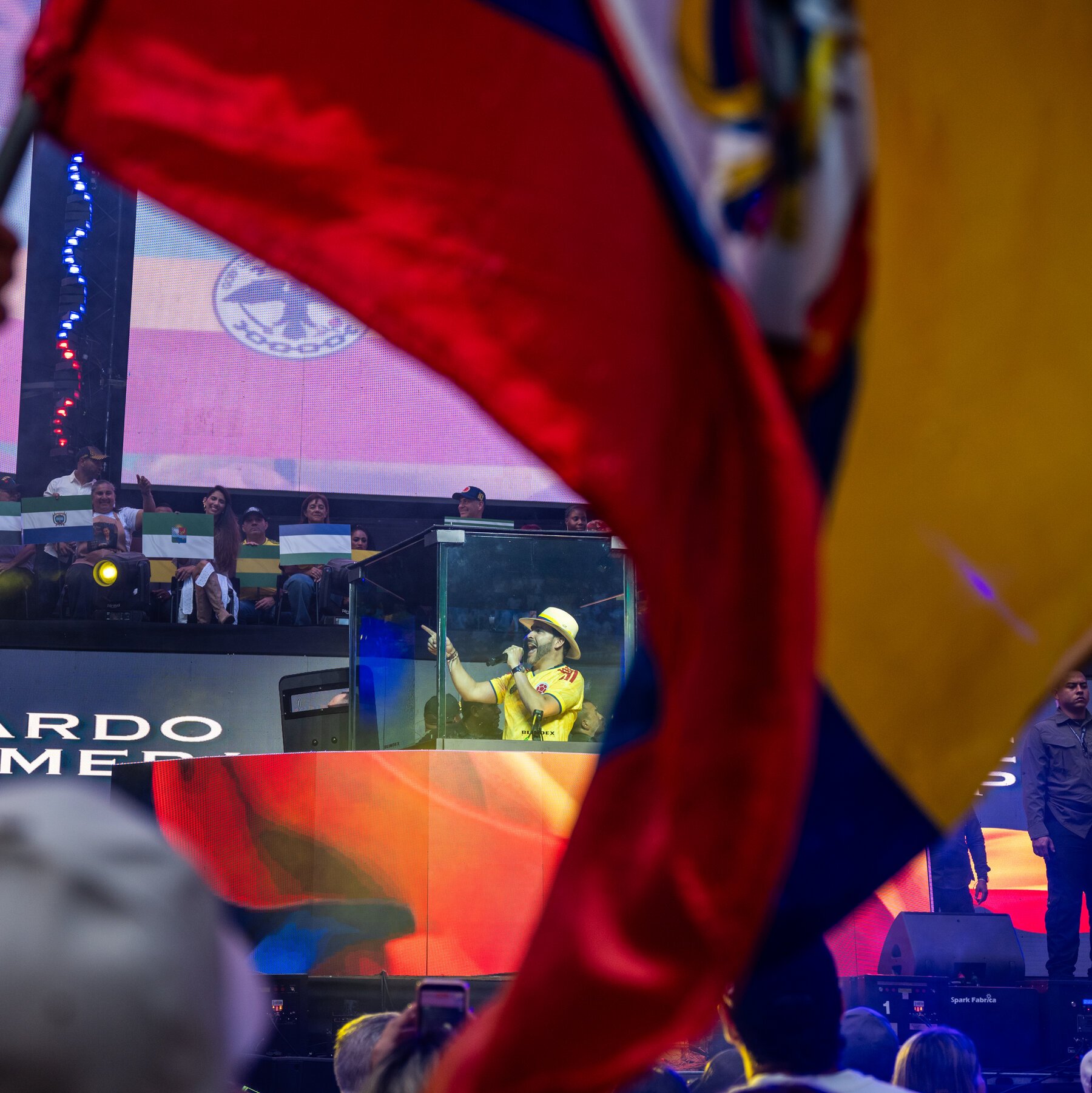 A man in a yellow shirt and hat sings into a microphone inside a glass booth at a stadium rally, with spectators waving flags above him.