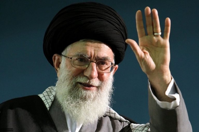 An elderly man with a white beard, wearing glasses and a black turban, raises his hand in a waving gesture against a dark background.
