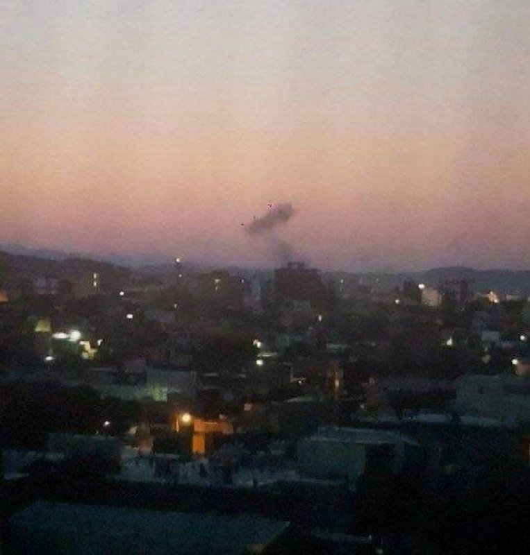 A plume of dark smoke rises above a hazy cityscape at dusk, with scattered lights visible among densely packed buildings.