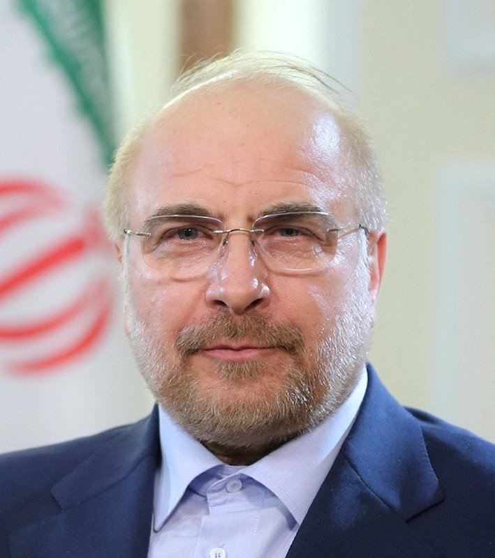A bearded, balding man wearing glasses and a blue suit smiles at the camera, with the Iranian flag visible behind him.