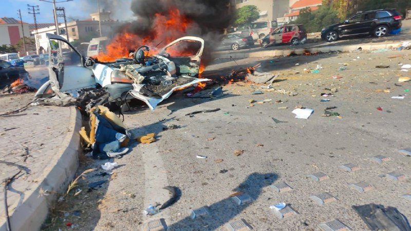 A white vehicle burns intensely on a debris-strewn street, with black smoke rising and damaged cars visible in the background.
