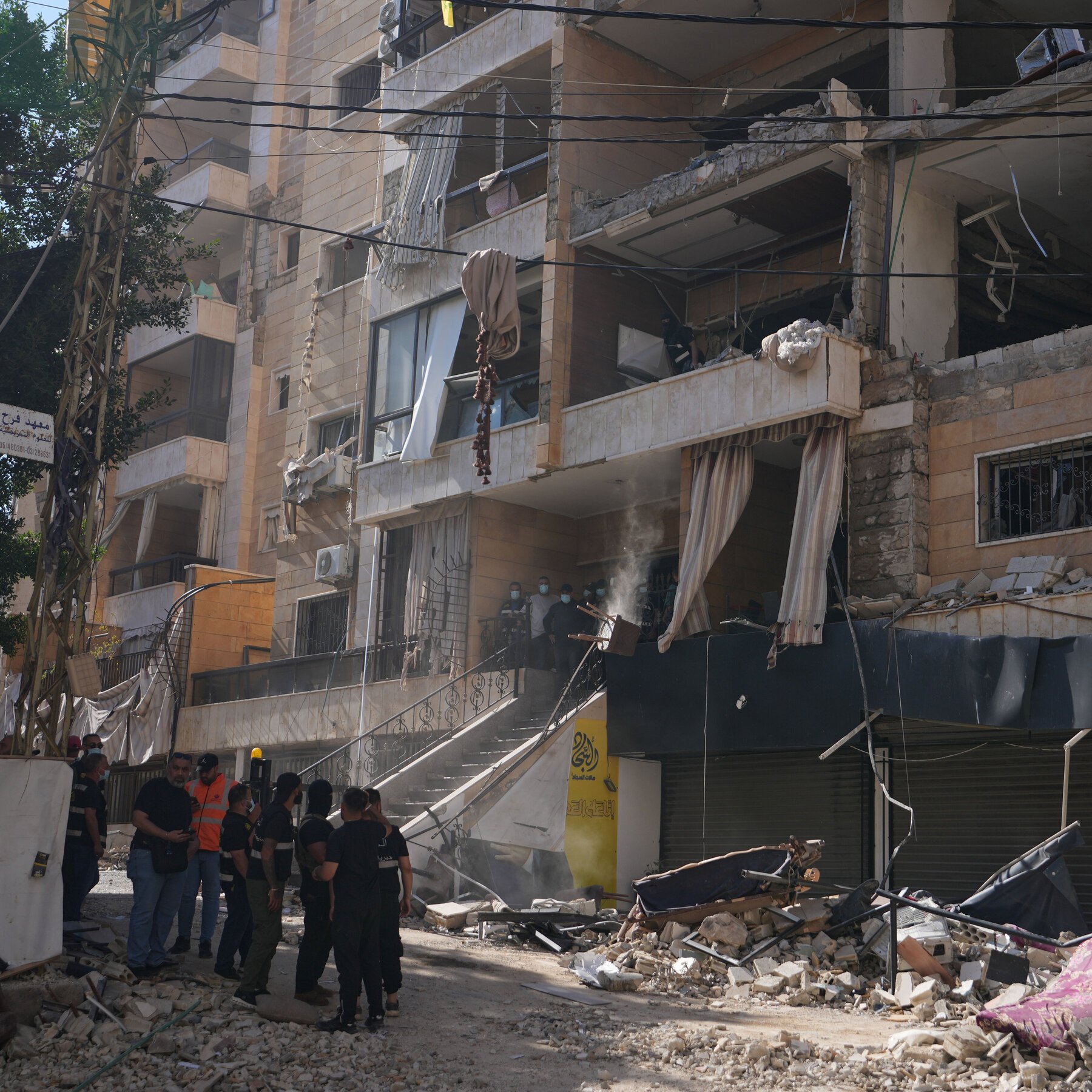 A multi-story residential building shows severe structural damage with blown-out balconies, hanging debris, and rubble scattered below as masked personnel inspect the site amid dust and smoke.