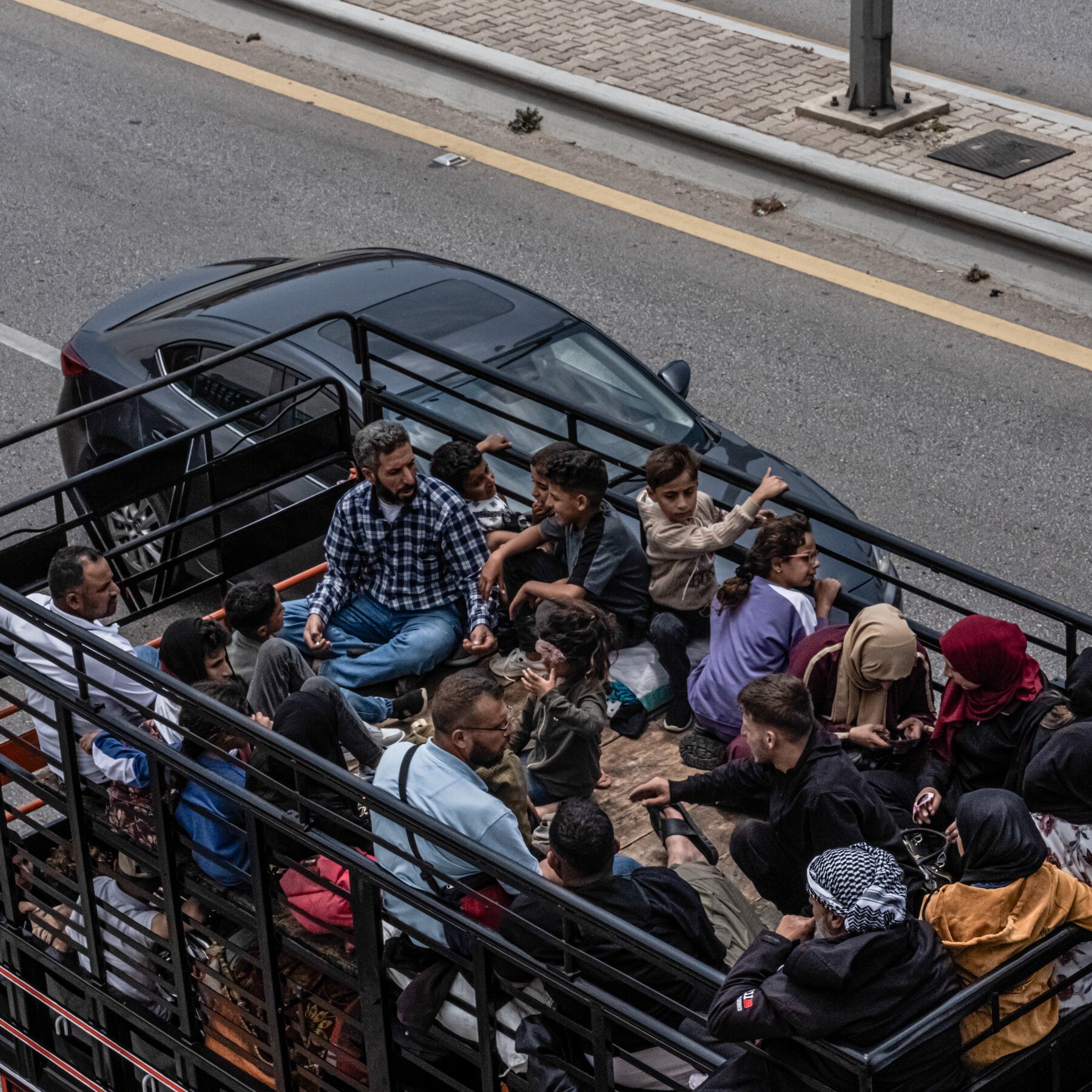 A crowded open-bed truck loaded with men, women, and children travels along a paved road next to a sidewalk.