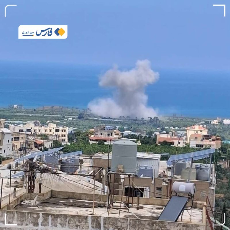 A plume of white smoke rises above a coastal town with rooftops, water tanks, and solar panels in the foreground, with the sea visible beyond.