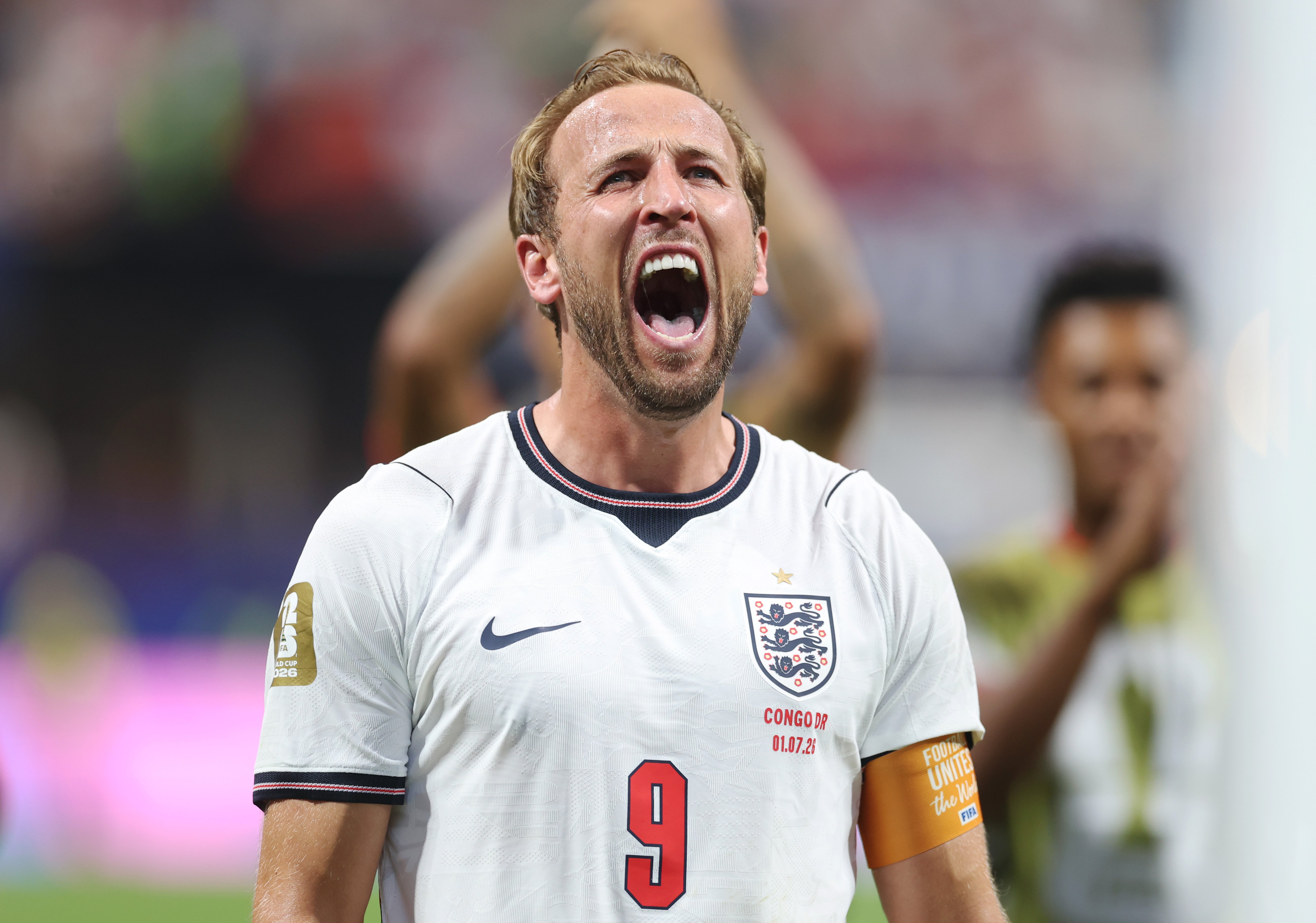 A blonde, bearded soccer player wearing a white England national team jersey with the number 9 shouts with his mouth wide open, displaying visible emotion during a match.