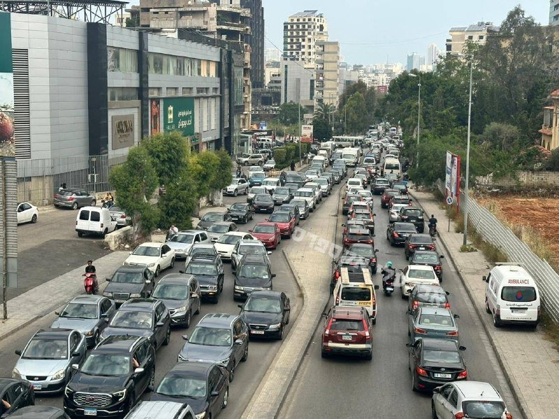 Heavy traffic congestion fills a multi-lane urban road lined with commercial buildings, trees, and an ambulance, with motorcyclists navigating between stationary and slow-moving cars.