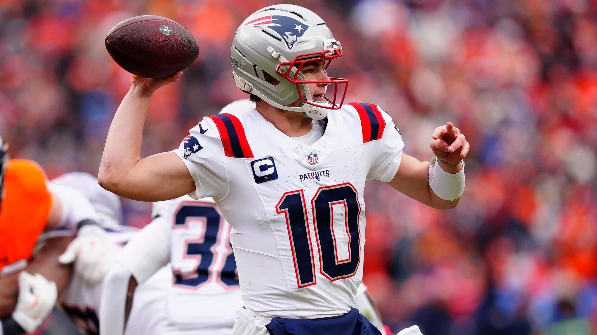 A New England Patriots quarterback wearing jersey number 10 winds up to throw a football during an NFL game, with an orange-clad crowd visible in the blurred background.