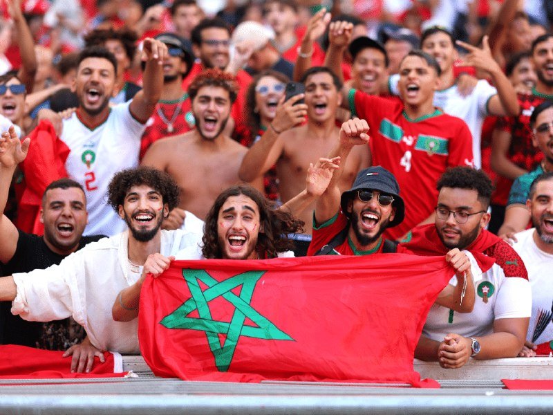 Cheering fans in red and white jerseys hold up a red flag with a green star while celebrating in stadium stands.